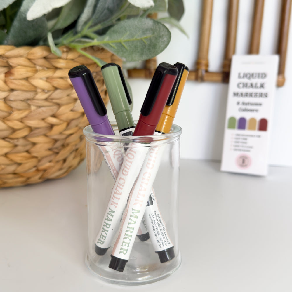 Coloured liquid chalk markers in red yellow purple and green styled in a glass jar with plants in the background